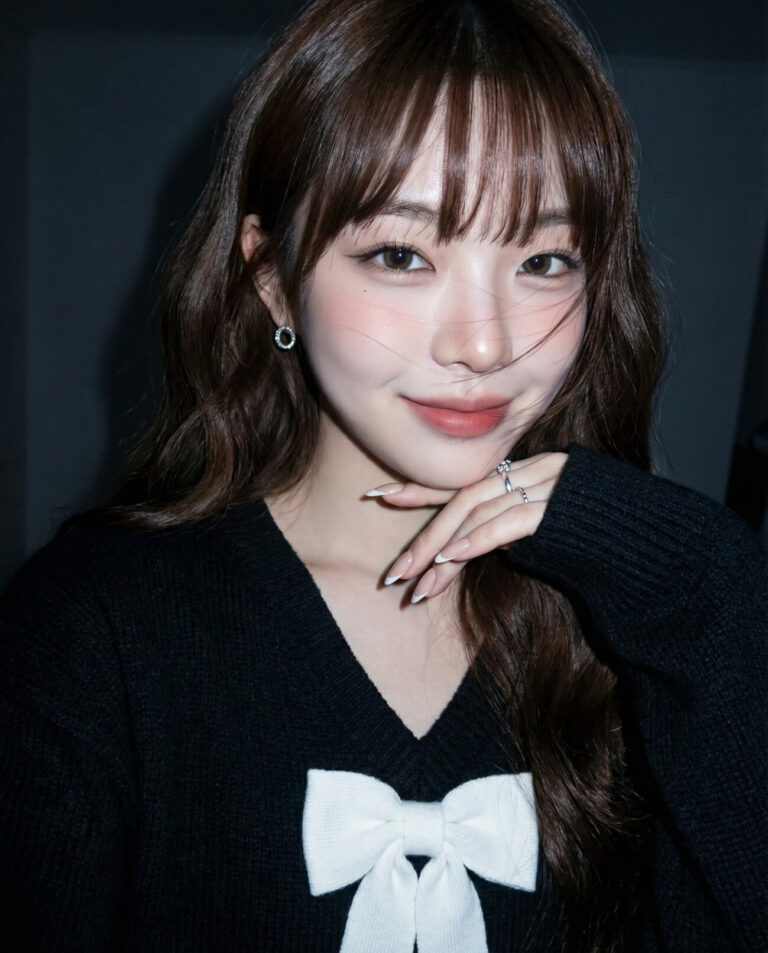 Gemini Ai Generated Image: a close-up portrait of a smiling young woman with long, wavy brown hair and bangs. She is wearing a black V-neck sweater accented with a prominent white bow at the center. Her hand is gently raised near her chin, displaying silver rings and a French manicure, while small hoop earrings complete the look. The lighting highlights her rosy cheeks and face against a dark background, creating a soft and stylish aesthetic.