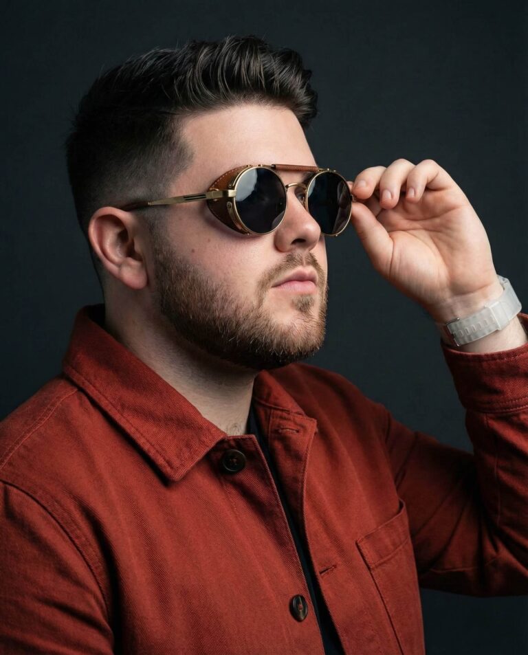Gemini Generated Image: a man with dark, styled hair and a short beard posing against a solid dark grey background. He is wearing a rust-colored jacket and distinctive round, black sunglasses with gold frames and side shields. With his hand, which sports a white watch, he is holding the edge of his glasses while looking off to the side.